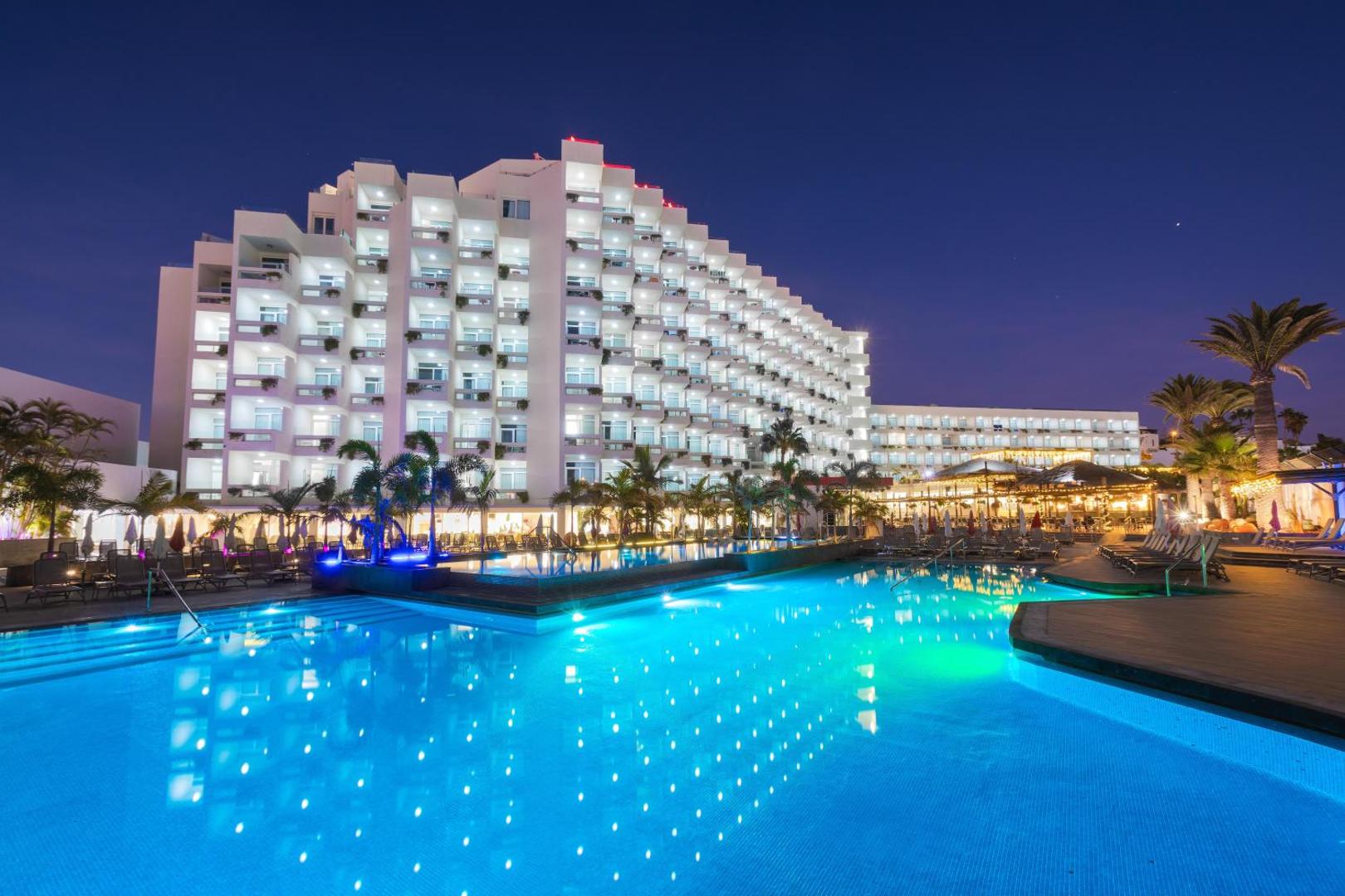 Alexandre Hotel Troya izgaismotais baseins un viesnīcas fasāde vakarā Tenerifē — zilas gaismas baseinā un mājīgs apgaismojums uz balkoniem.