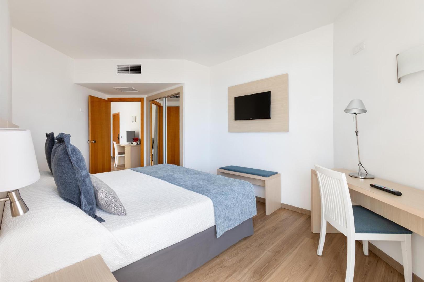 Alexandre Hotel Troya gaišs un moderns divvietīgs numurs Tenerifē ar koka grīdu, rakstāmgaldu, sienas televizoru un ziliem akcentiem interjerā.