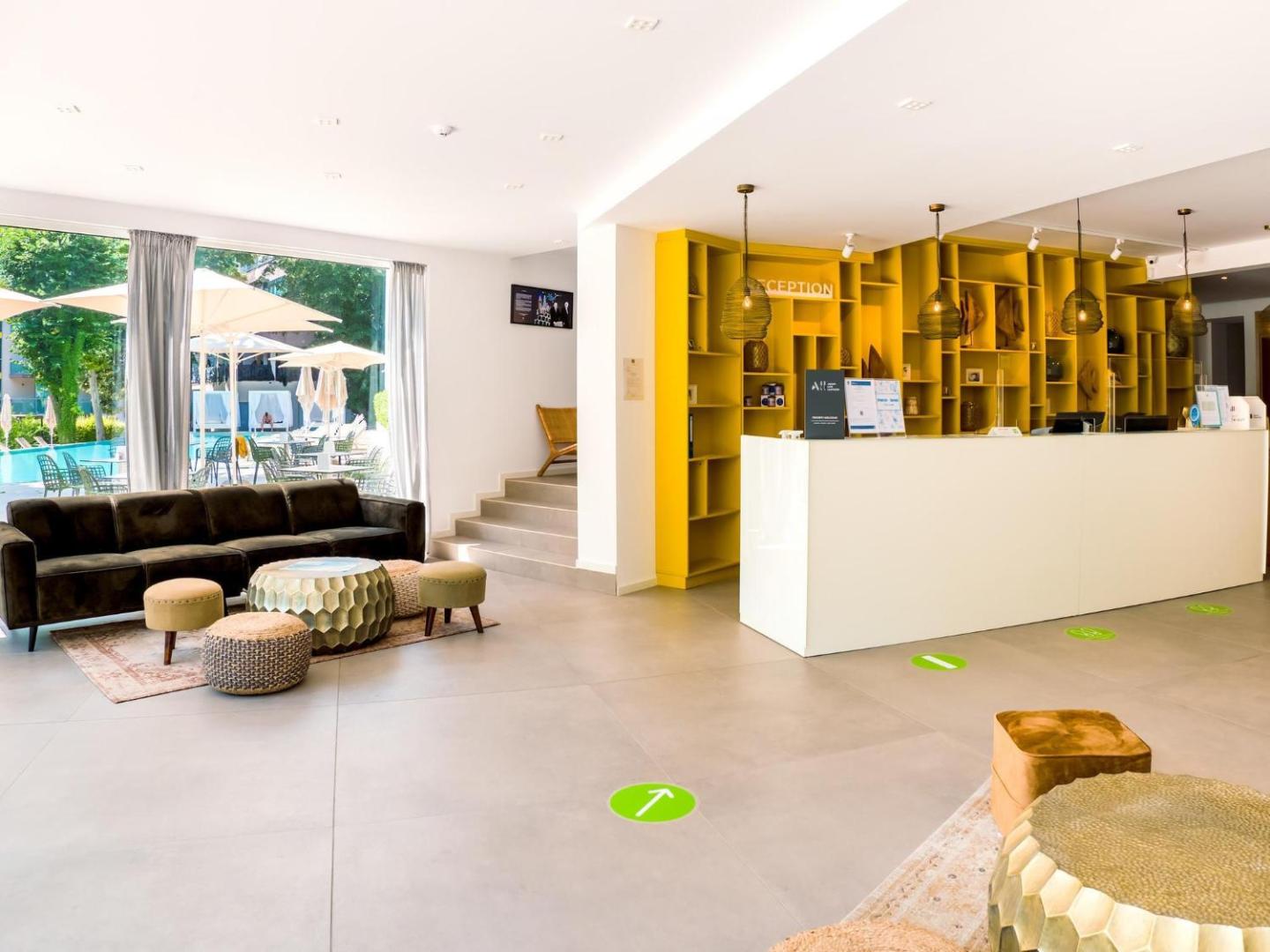 Ibis Styles Golden Sands Roomer Hotel gaišā recepcija ar dzelteniem plauktiem un skatu uz baseina zonu