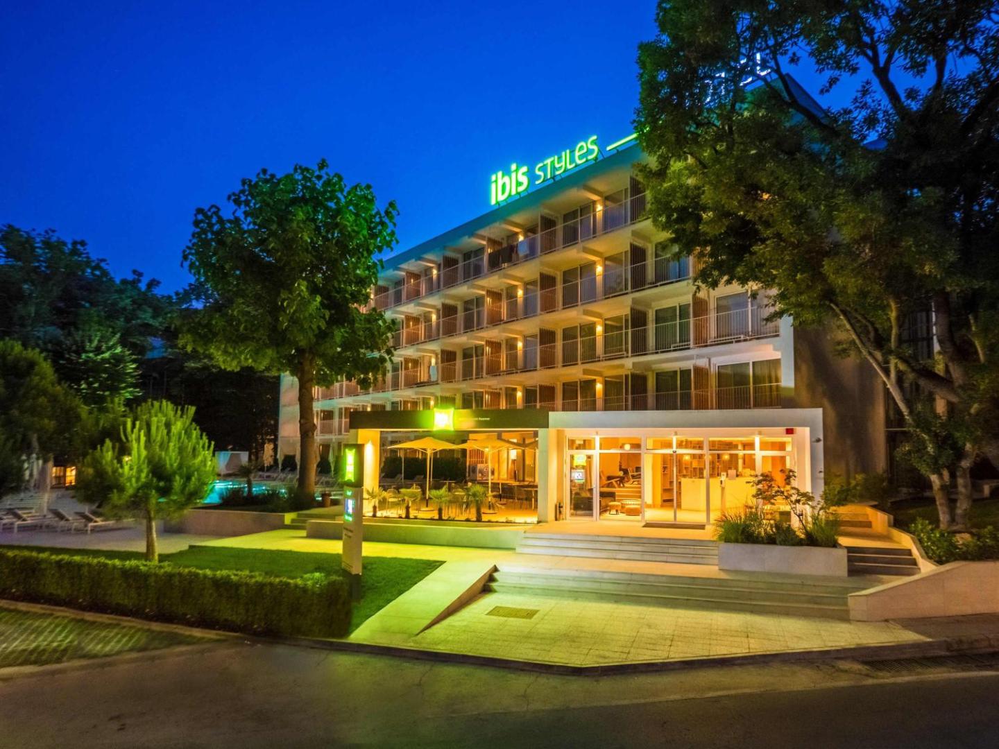 Ibis Styles Golden Sands Roomer viesnīcas ēka vakarā ar izgaismotu logo un ieeju kūrortā Zelta smiltis, Bulgārijā