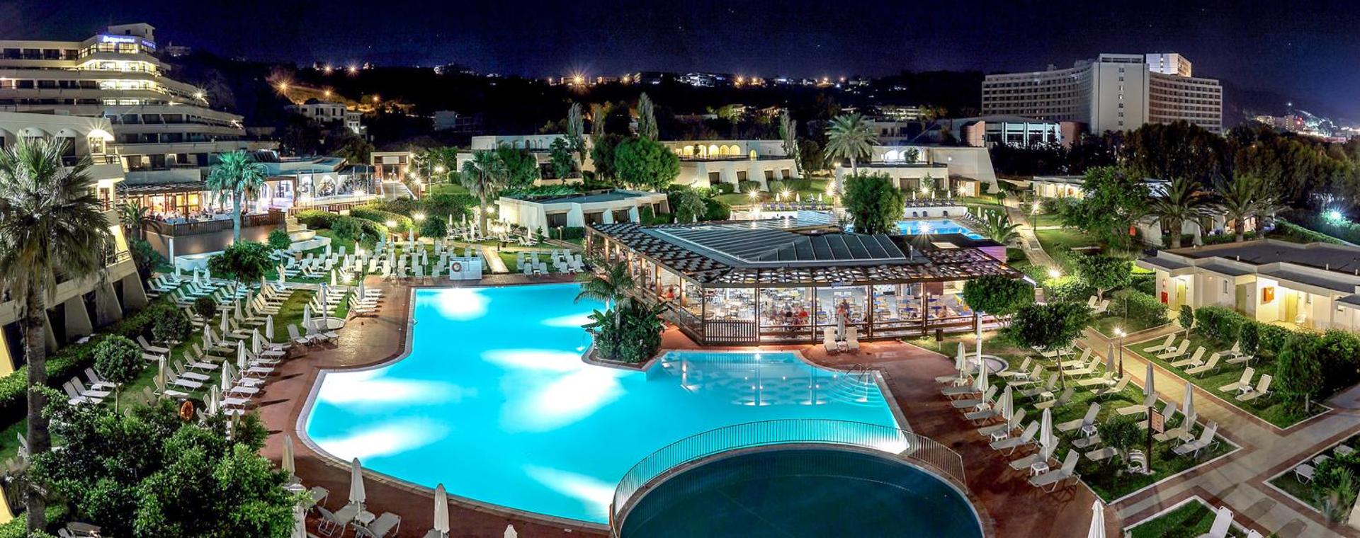 Sol by Melia Cosmopolitan Rhodes viesnīcas teritorija naktī ar izgaismotiem baseiniem, restorānu un skatu uz Rodas pilsētas gaismām