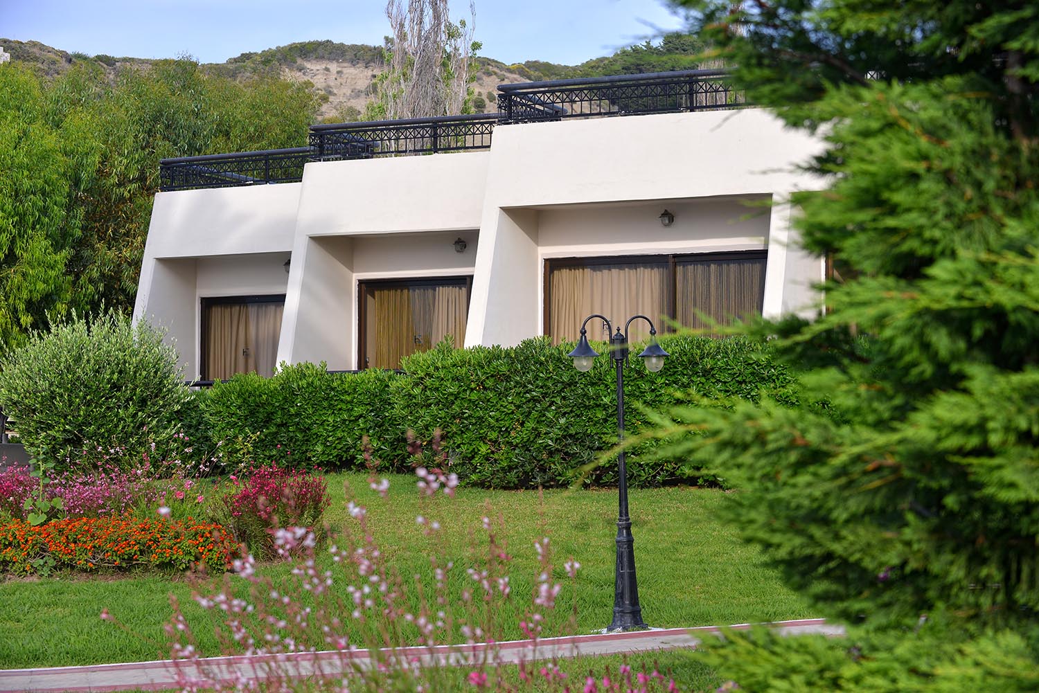 Sol by Melia Cosmopolitan Rhodes gaišie bungalo Rodas salā, ieskauti zaļos apstādījumos un ziedošās puķēs pie gājēju celiņa