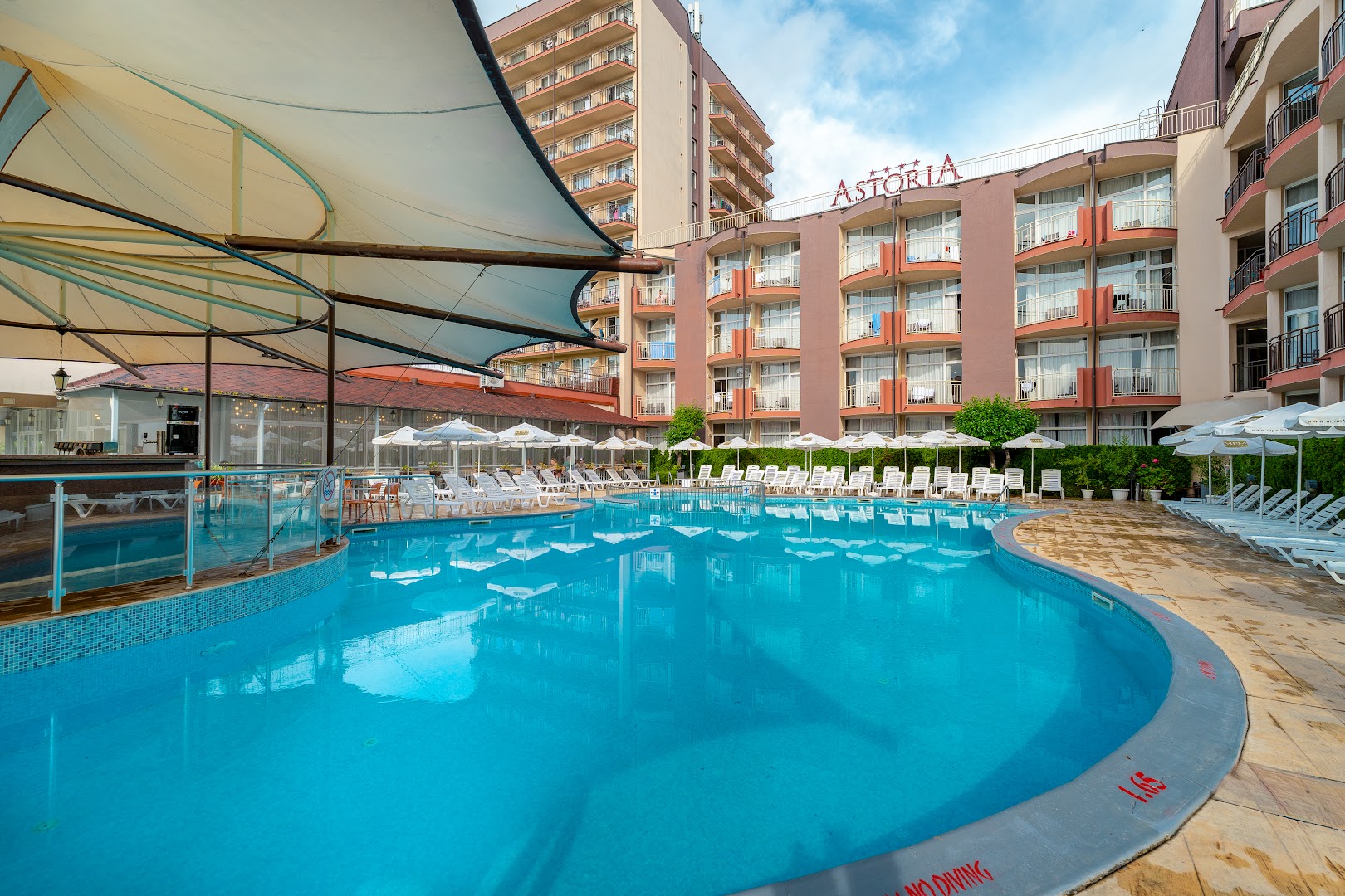 MPM Hotel Astoria viesnīcas ēka un lielais āra baseins Saulainajā krastā — saulessargi, sauļošanās krēsli un baseina bārs zem nojumes