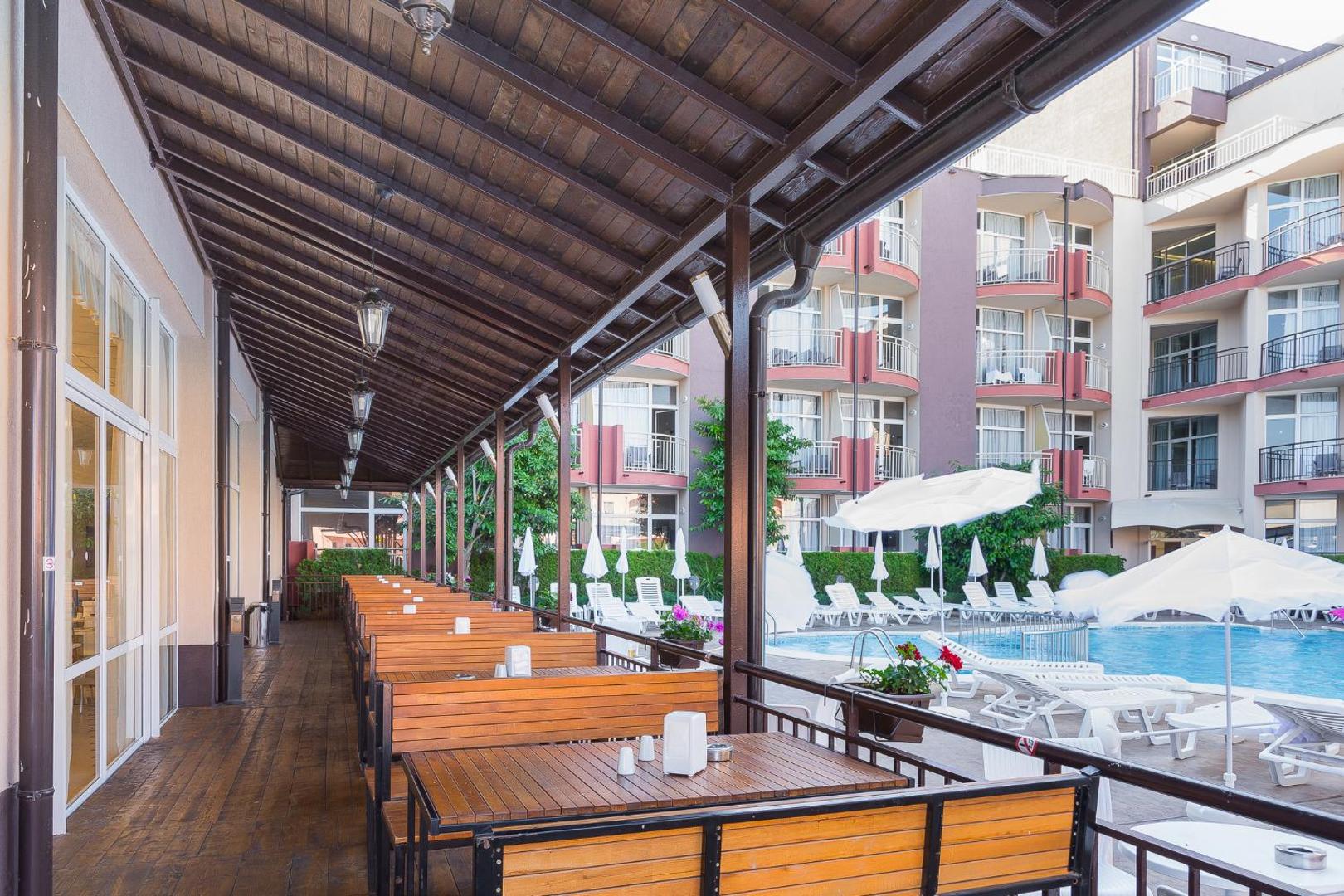 MPM Hotel Astoria āra terase ar koka galdiem un soliem pie baseina, fonā redzama viesnīcas ēka ar balkoniem
