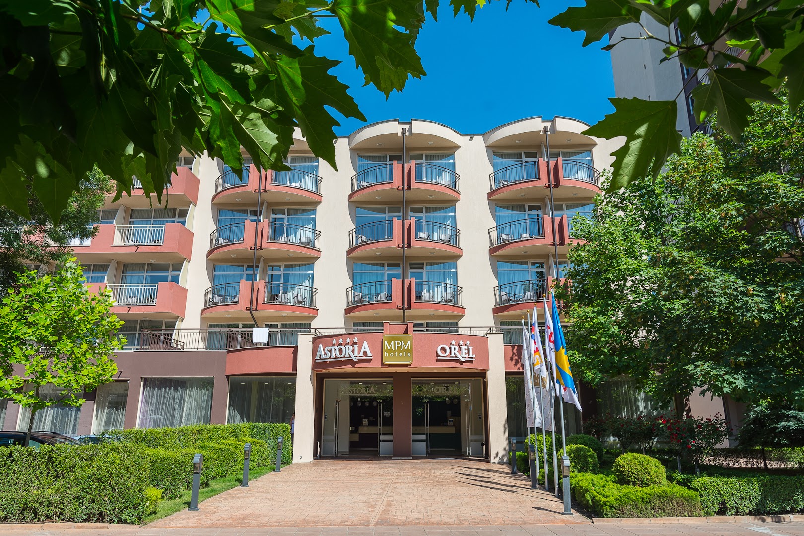 MPM Hotel Astoria un Orel viesnīcu galvenā ieeja ar karogiem un zaļiem apstādījumiem Saulainajā krastā