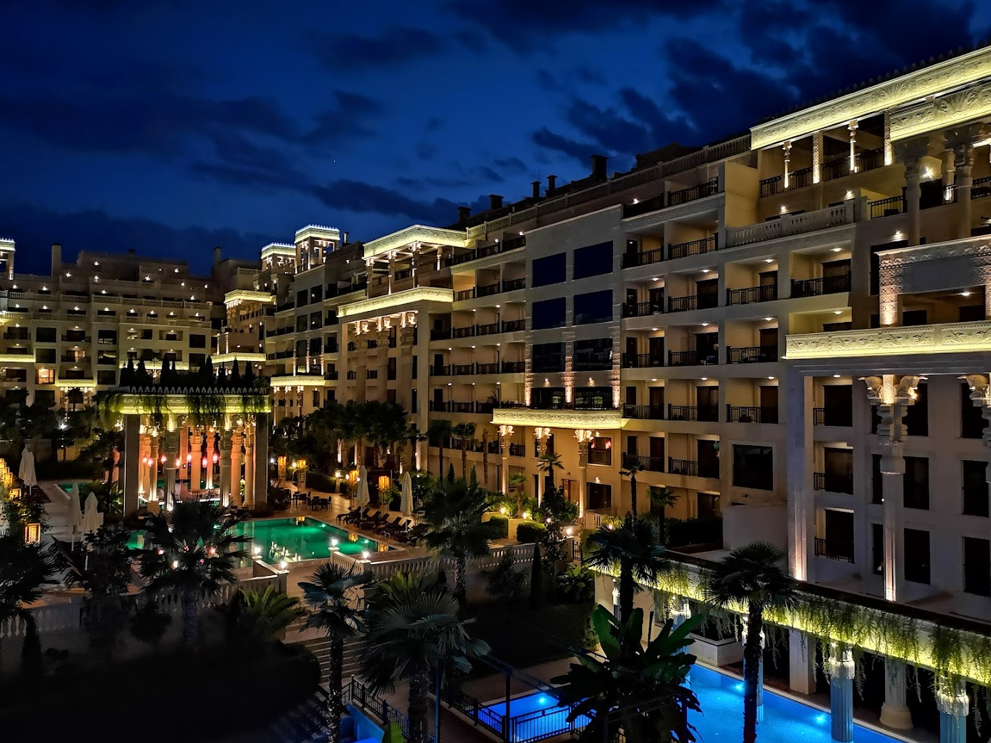 Skats uz Argisht Partez Hotel iekšpagalmu un baseiniem naktī ar dekoratīvo apgaismojumu un palmu siluetiem