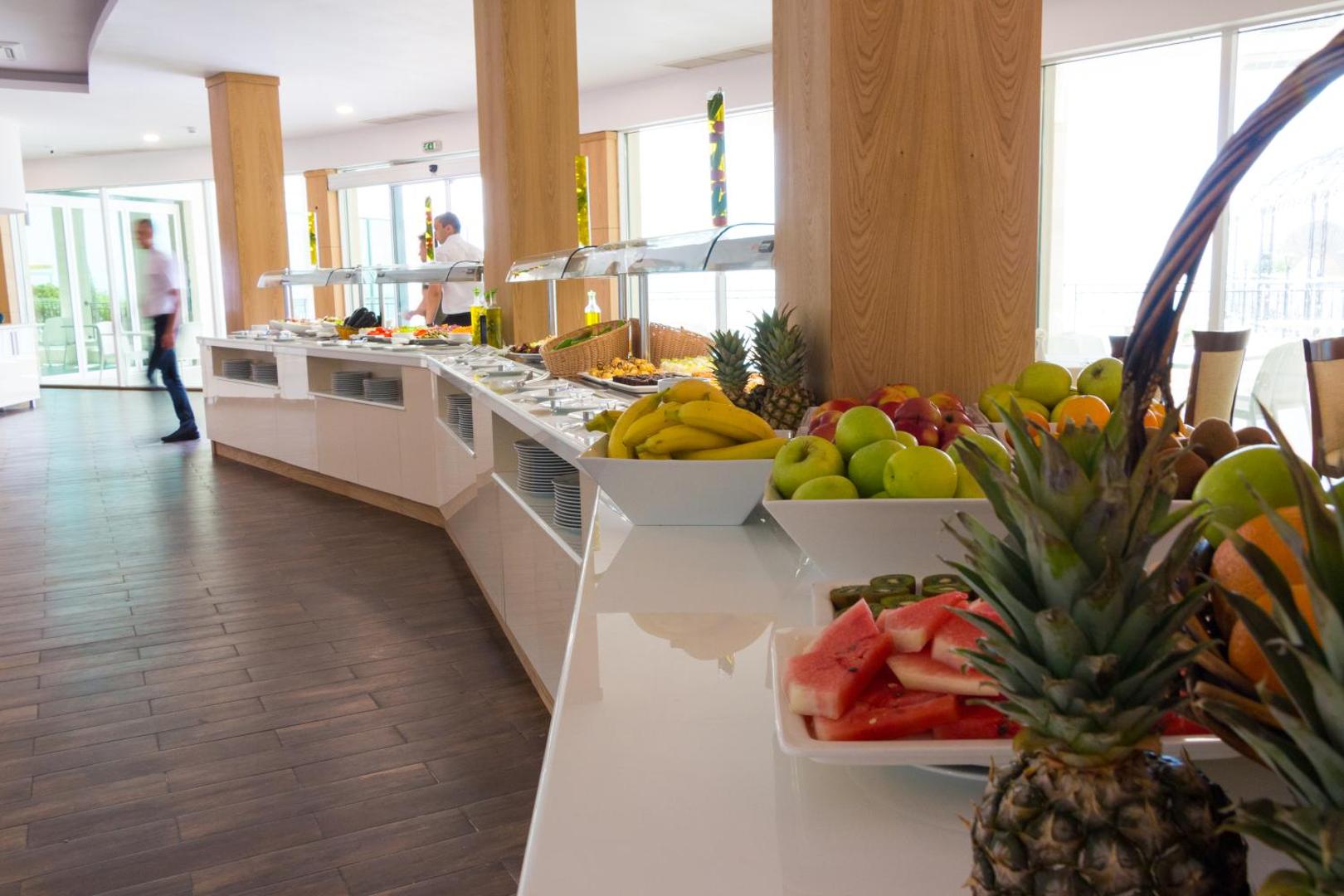 Bilyana Beach Hotel restorāna bufetes zona ar svaigiem augļiem, ananasiem un arbūziem — gaišs interjers ar koka elementiem
