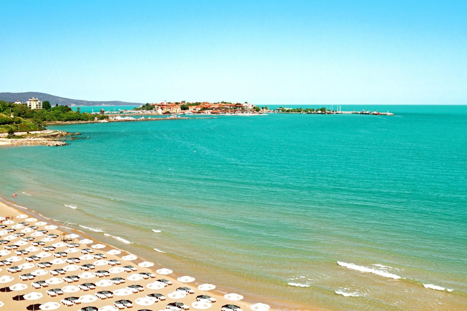 Panorāmas skats no Bilyana Beach Hotel uz Nesebru un Melno jūru — plaša smilšu pludmale ar baltiem saulessargiem