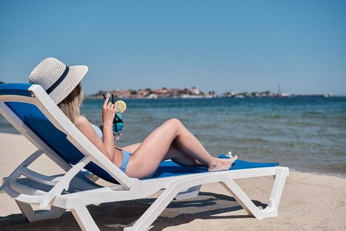 Viesis atpūšas sauļošanās krēslā Bilyana Beach Hotel pludmalē ar kokteili rokās — fona redzama Nesebāras vecpilsētas panorāma