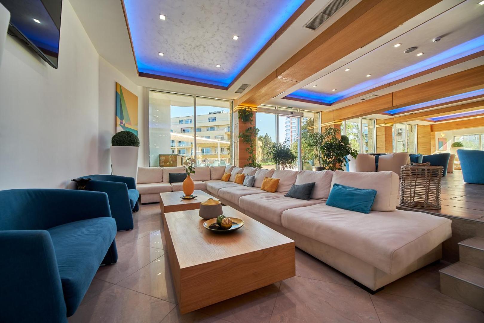 Bilyana Beach Hotel lobby bārs un atpūtas zona ar gaišiem dīvāniem, ziliem akcentiem un plašiem logiem uz kūrorta teritoriju