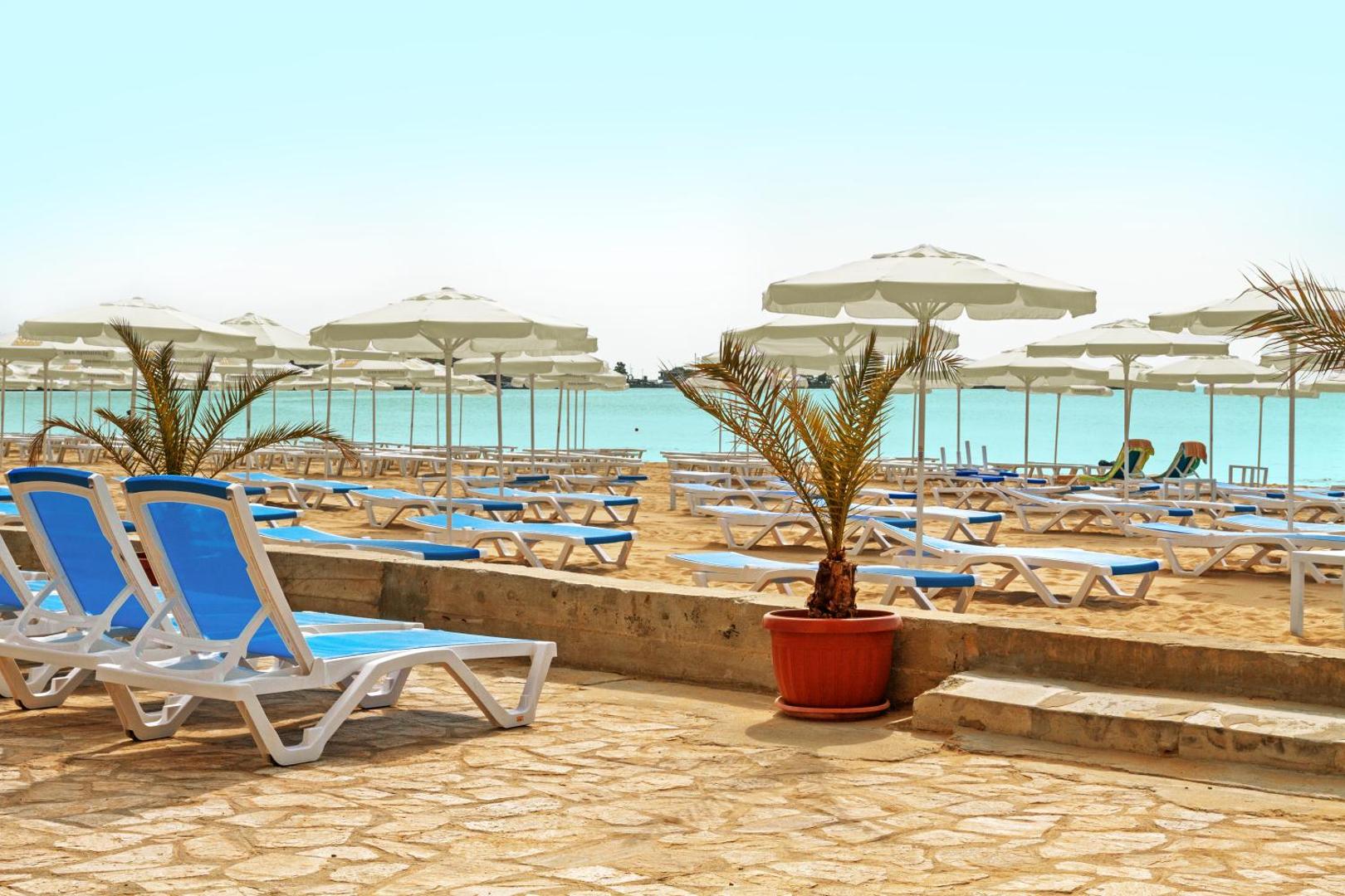 Bilyana Beach Hotel privātā pludmale Nesebārā ar ziliem sauļošanās krēsliem un baltiem saulessargiem pie dzidra ūdens