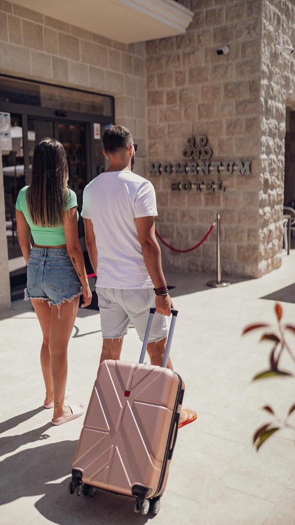 Pāris ar ceļojuma koferi ierodas Momentum by Aycon boutique viesnīcā — ieeja ar akmens apdari un viesnīcas logo