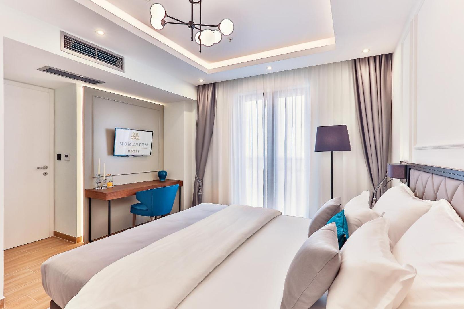 Skats uz Momentum by Aycon Boutique Hotel numuru ar TV zonu, rakstāmgaldu un gaišiem aizkariem pie balkona durvīm