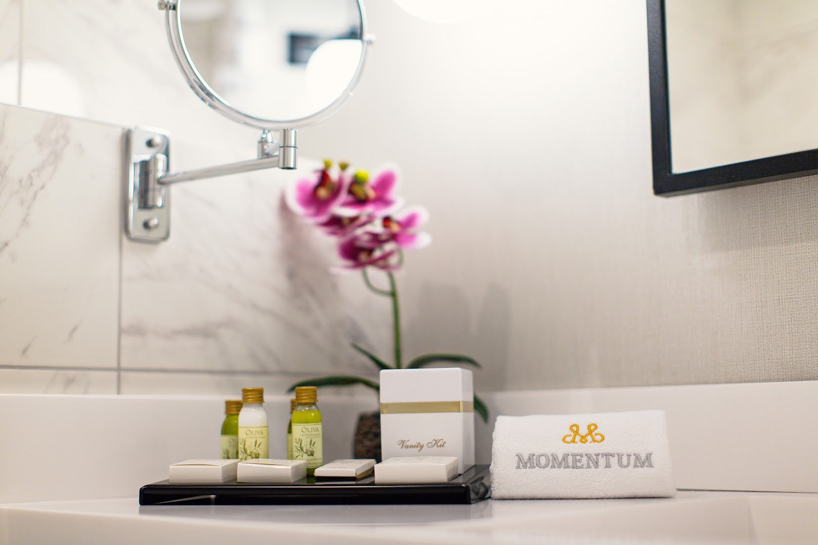 Tuvplāns ar Momentum by Aycon Boutique Hotel bezmaksas kosmētikas piederumiem, orhideju un zīmola dvieli uz balta vannas istabas virsmas.