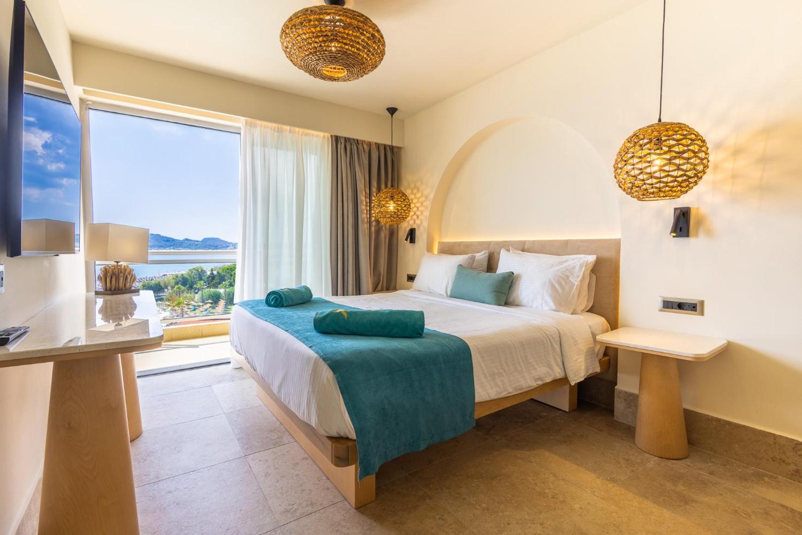 Pegasos Beach Hotel moderns divvietīgs numurs ar gaišu interjeru, pītiem gaismekļiem un balkonu, no kura paveras skats uz jūru.