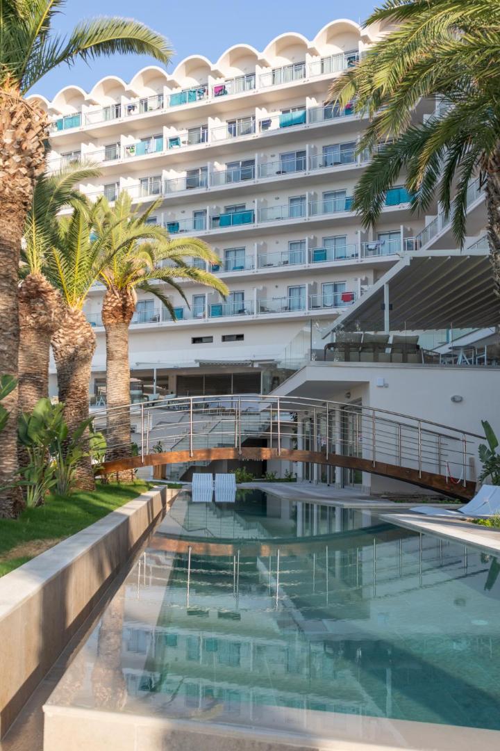 Pegasos Beach Hotel Rodā — skats uz baseinu ar tiltiņu un viesnīcas numuru balkoniem dienas gaismā