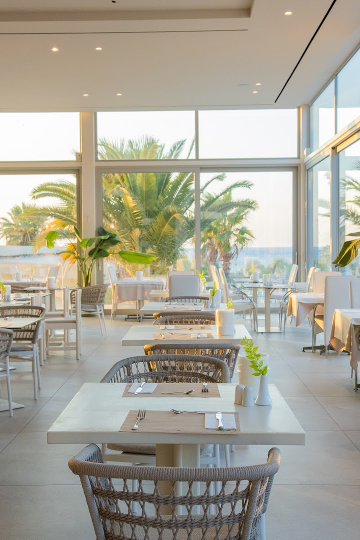 Gaišs Pegasos Beach Hotel & Resort restorāns ar panorāmas logiem, skatu uz palmām un jūru Rodas piekrastē.
