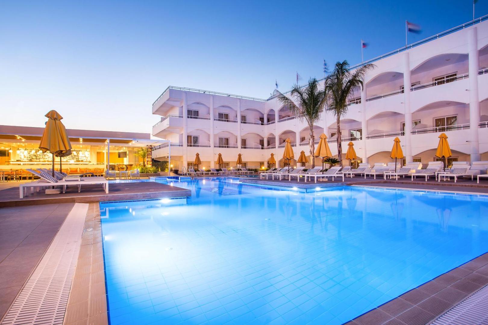 Orion Hotel Rhodes izgaismots āra baseins vakarā ar viesnīcas korpusu un bāru fonā Faliraki kūrortā, Rodā.