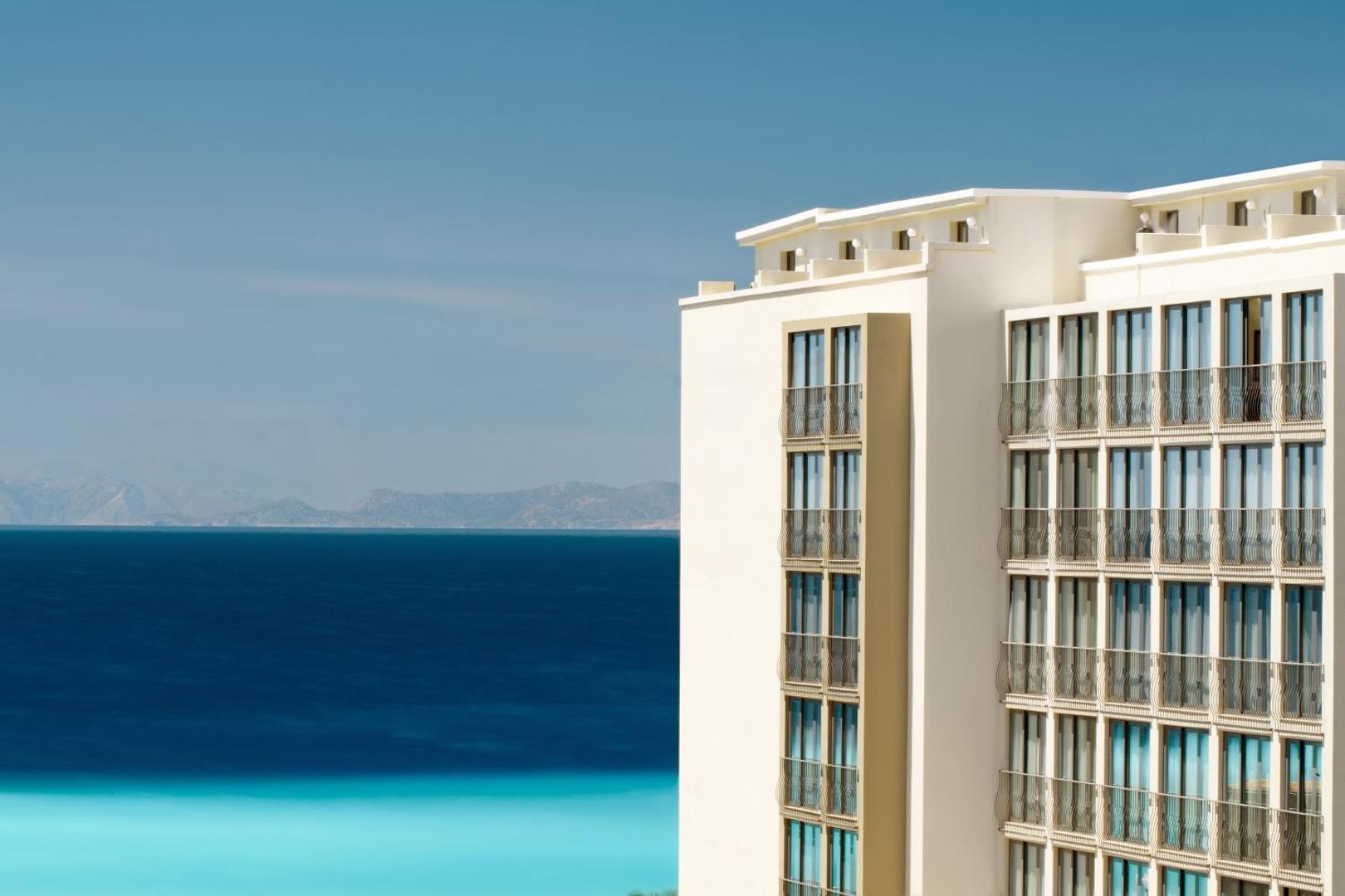 Mitsis La Vita Beach Hotel ēkas stūris uz koši zilas Egejas jūras un kalnu fona Rodas salā