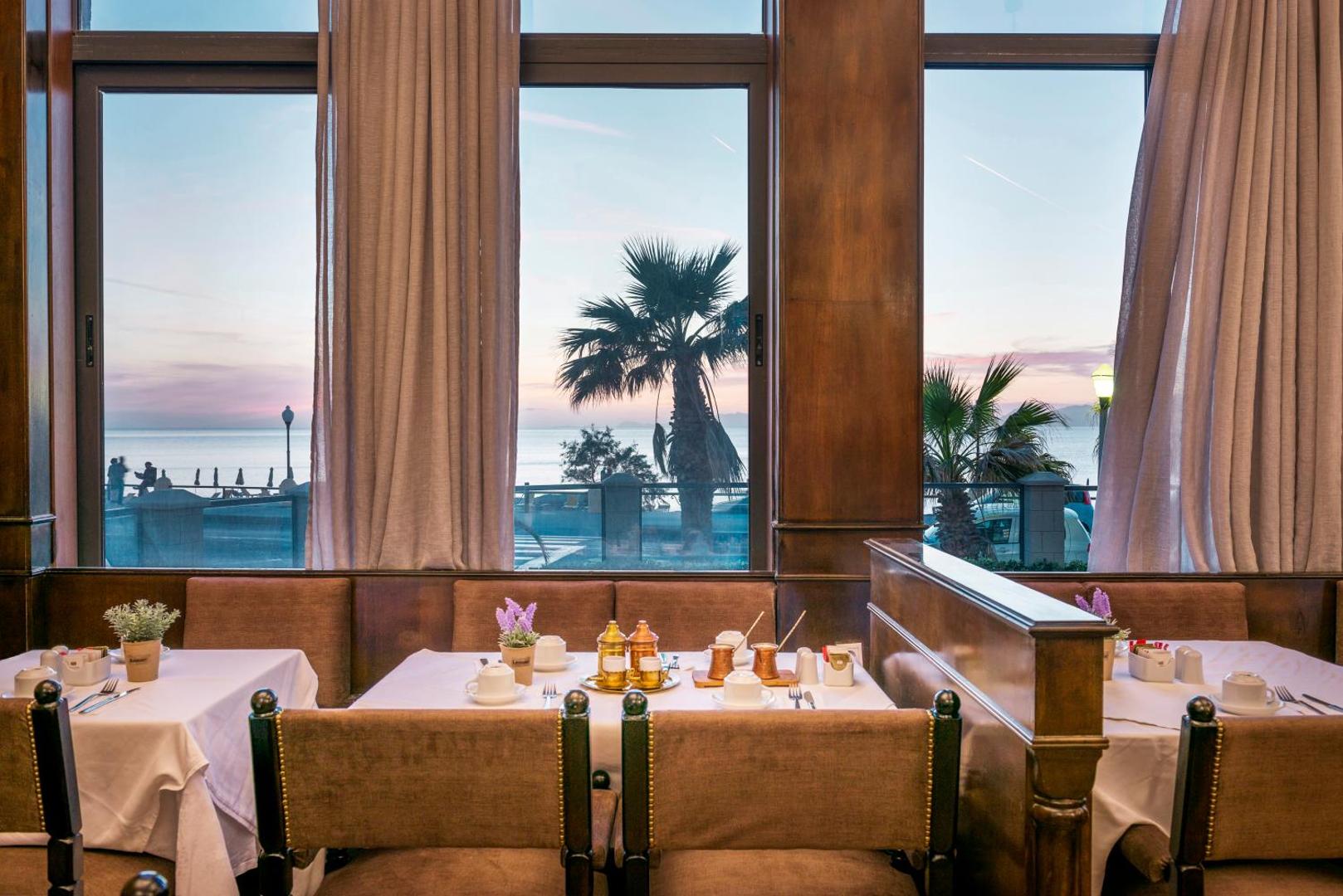 Mitsis La Vita Beach Hotel restorāna skats caur logu uz jūru un palmām saulrietā Rodas pilsētā, Grieķijā