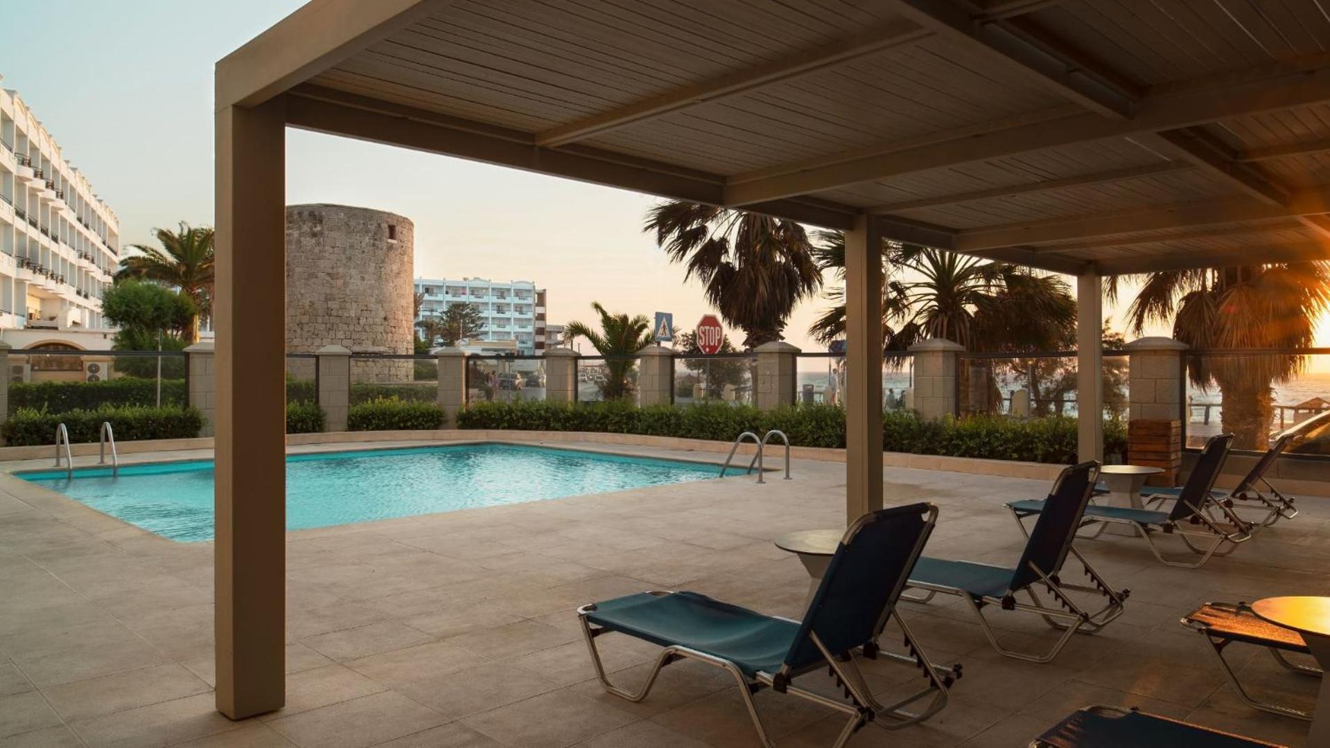 Mitsis La Vita Beach Hotel atpūtas zona pie baseina ar sauļošanās krēsliem zem nojumes vakara gaismā