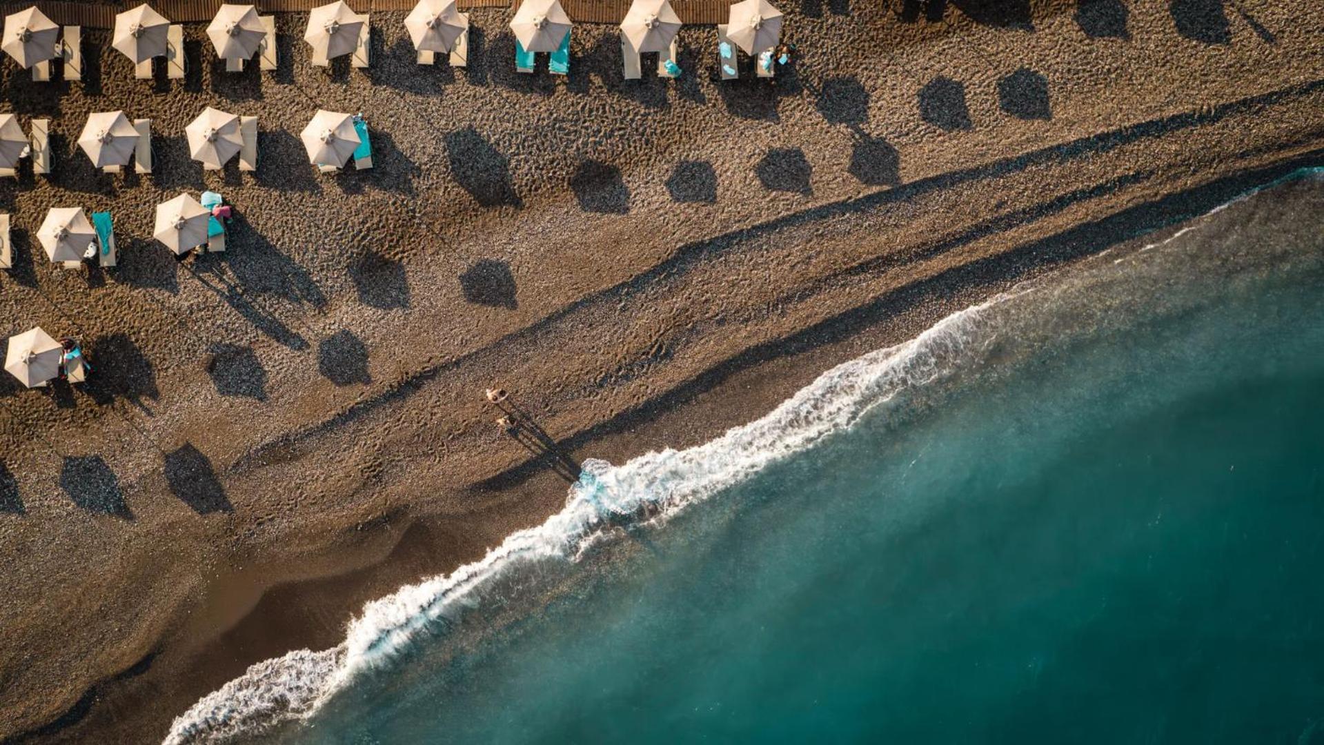 Aerofoto skats uz Mitsis La Vita Beach Hotel oļu pludmali ar saulessargiem un dzidru tirkīza ūdeni