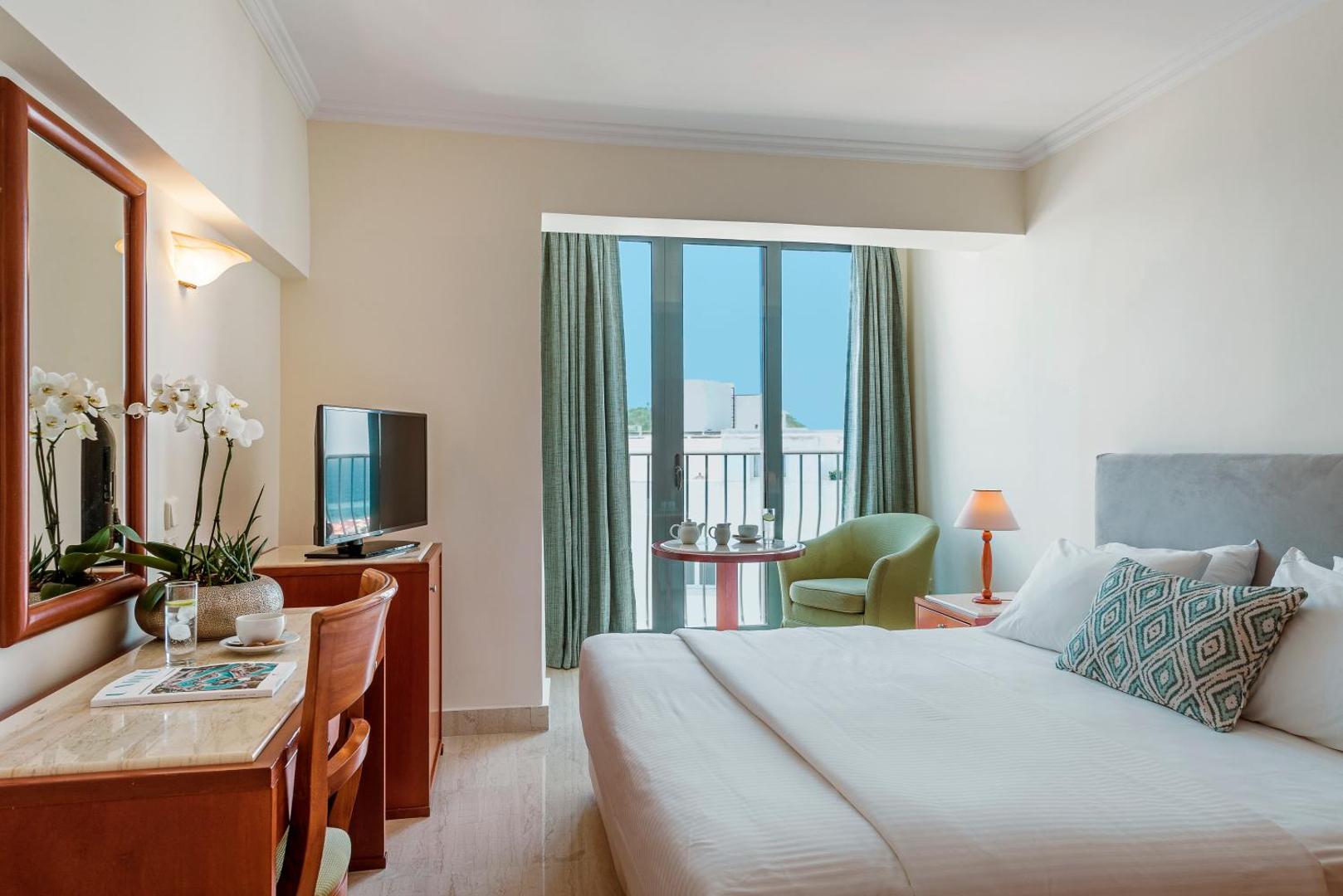 Gaišs Mitsis La Vita Beach Hotel viesnīcas numurs ar divguļamo gultu, rakstāmgaldu, televizoru un atpūtas krēslu pie loga