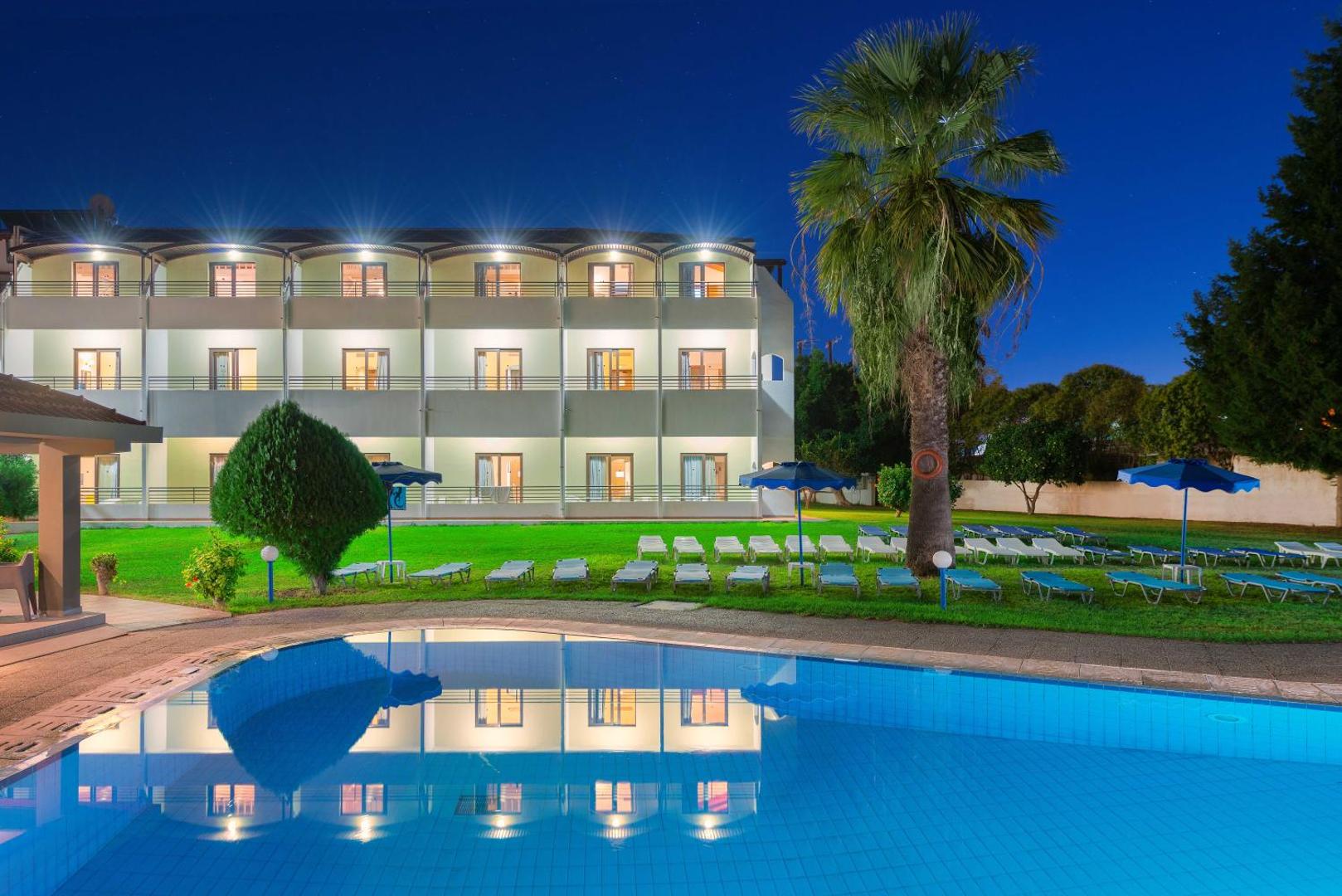 Matoula Beach Hotel fasāde un āra baseins vakarā Rodas salā — izgaismoti numuri ar balkoniem un atpūtas krēsli zālienā pie palmas
