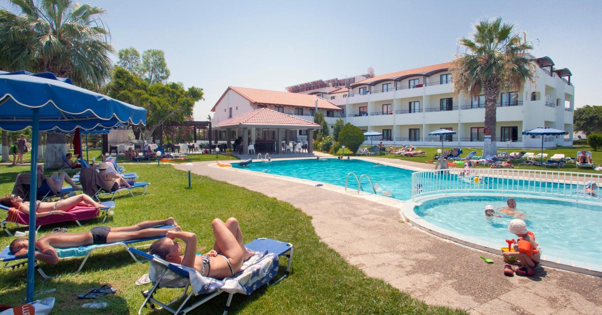Matoula Beach Hotel galvenais baseins un bērnu baseins saulainā dienā — viesi atpūšas sauļošanās krēslos zālienā pie viesnīcas ēkas