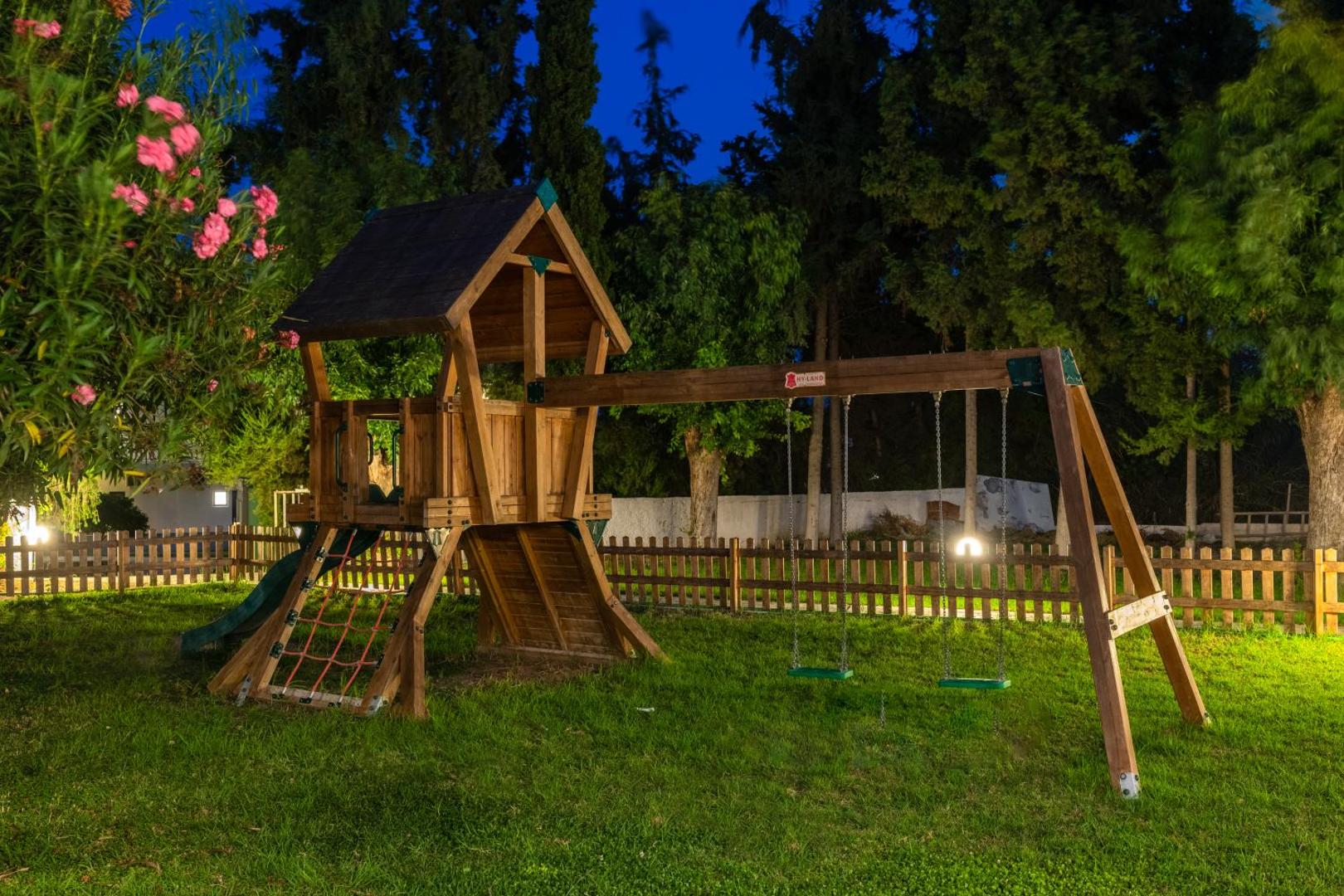 Matoula Beach Hotel koka rotaļu laukums vakarā Rodas salā — apgaismotas šūpoles un slidkalniņš bērniem zaļā dārzā pie koka sētas