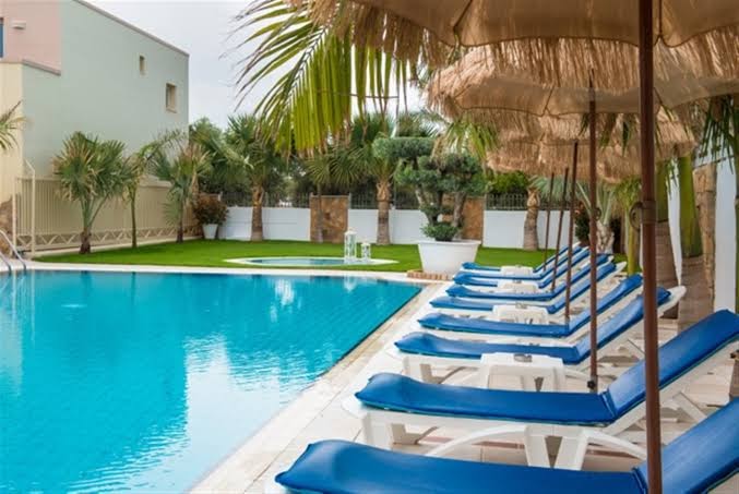 Sauļošanās krēsli ar saulessargiem pie Danaides Apartments baseina — palmas un zaļš zāliens viesnīcas teritorijā