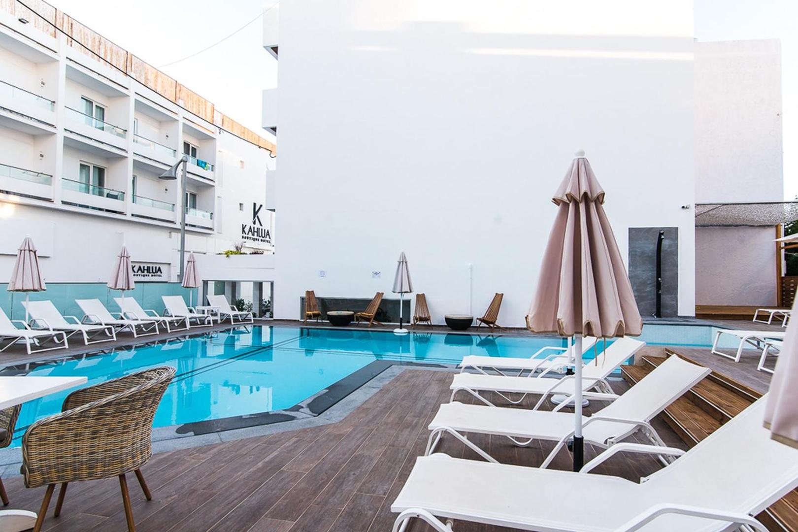 Kahlua Boutique Hotel baseina zona ar koka terasi un baltiem zviļņiem Krētā — fonā redzama viesnīcas modernā fasāde un saulessargi