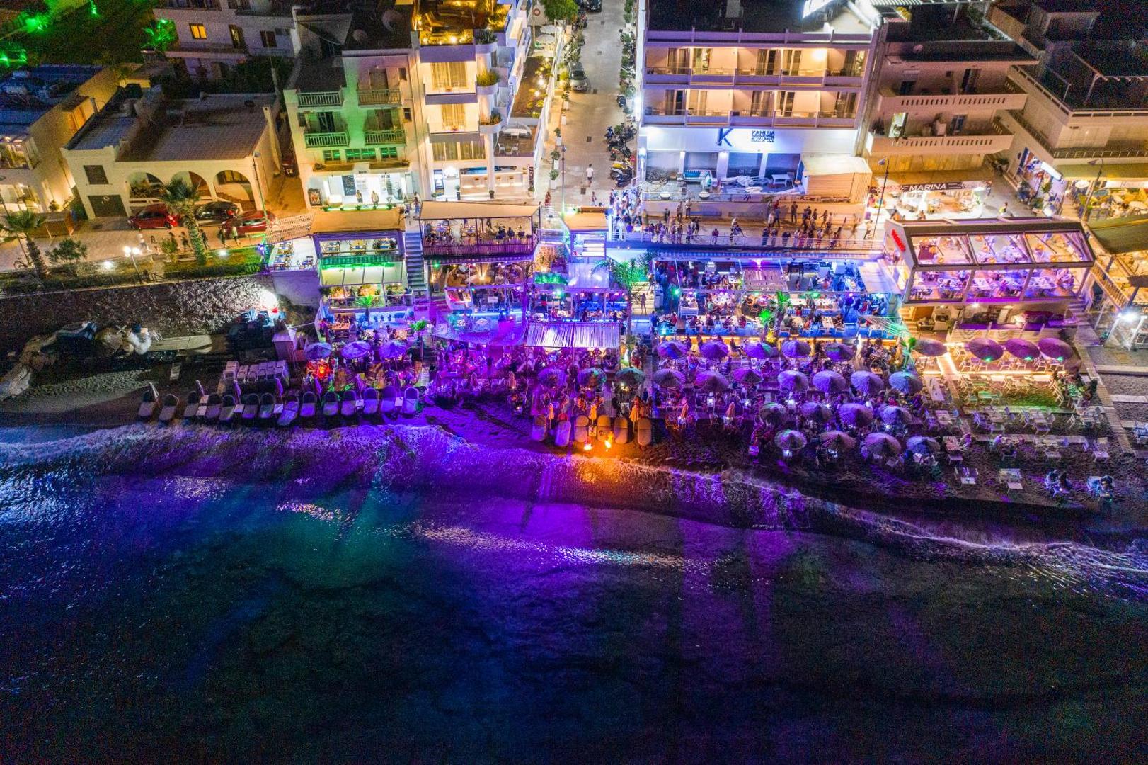 Kahlua Boutique Hotel skats no putna lidojuma naktī Hersonisā, Krētā — izgaismota pludmale, restorāni un piekrastes promenāde ar atpūtas zonām