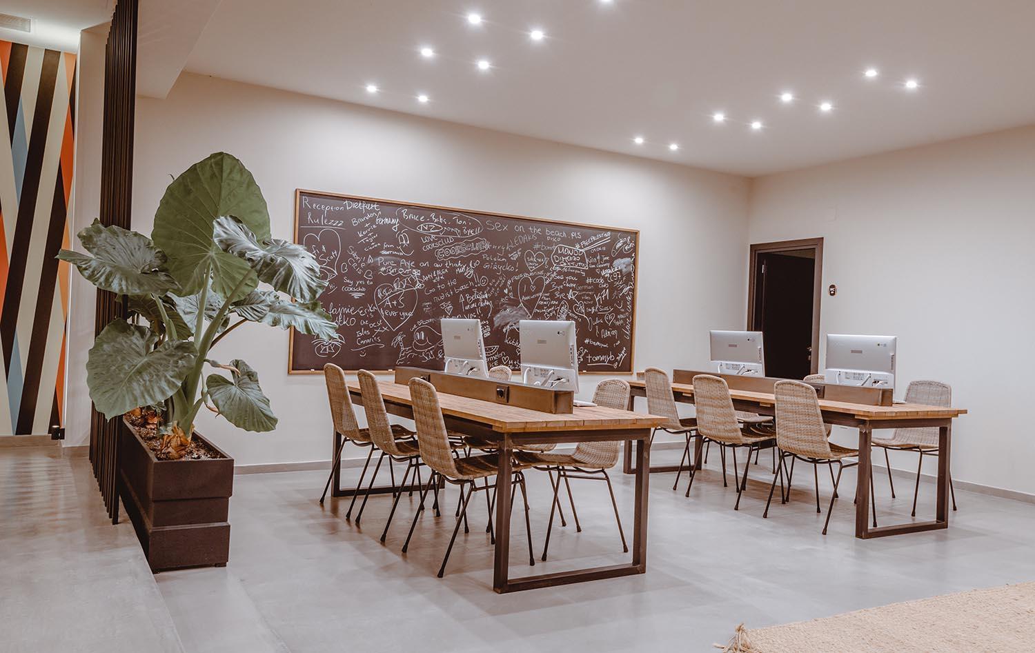 Cook's Club Hersonissos kopstrādes telpa ar datoriem, koka galdiem un lielu tāfeli pie sienas — moderna vide darbam un atpūtai