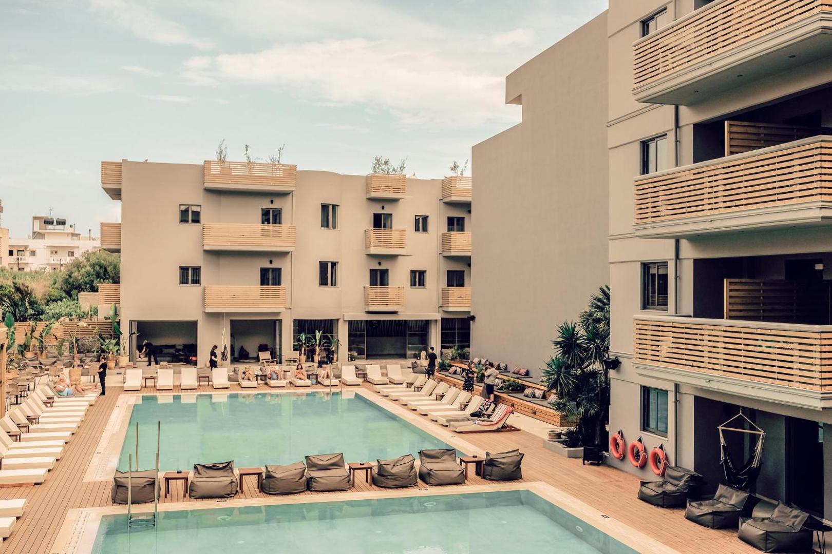 Cook's Club Hersonissos viesnīcas centrālais baseins Hersonisā, Krētā — atpūtas zona ar sauļošanās krēsliem, sēžammaisiem un modernu korpusu arhitektūru fonā.
