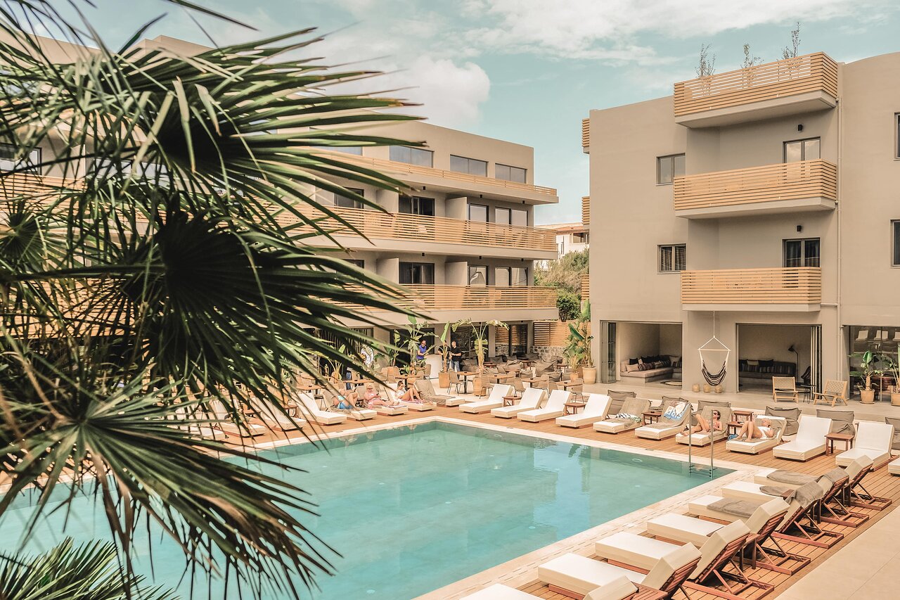 Cook's Club Hersonissos viesnīcas pagalms ar baseinu un modernām ēkām Hersonisā — sauļošanās zona ar ērtiem zviļņiem un palmām