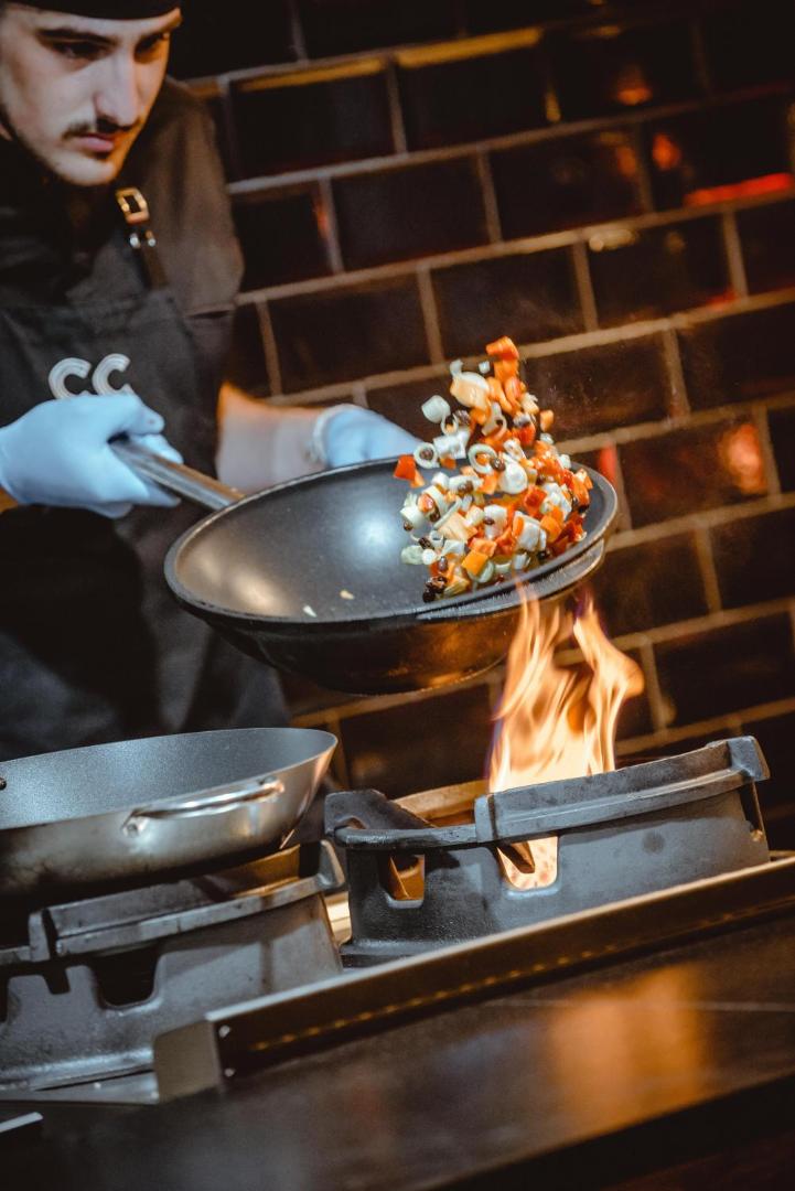 Cook's Club Hersonissos pavārs gatavo dārzeņus vokpannā uz atklātas uguns — dinamiska kulinārijas šova elements viesnīcas restorānā Krētā