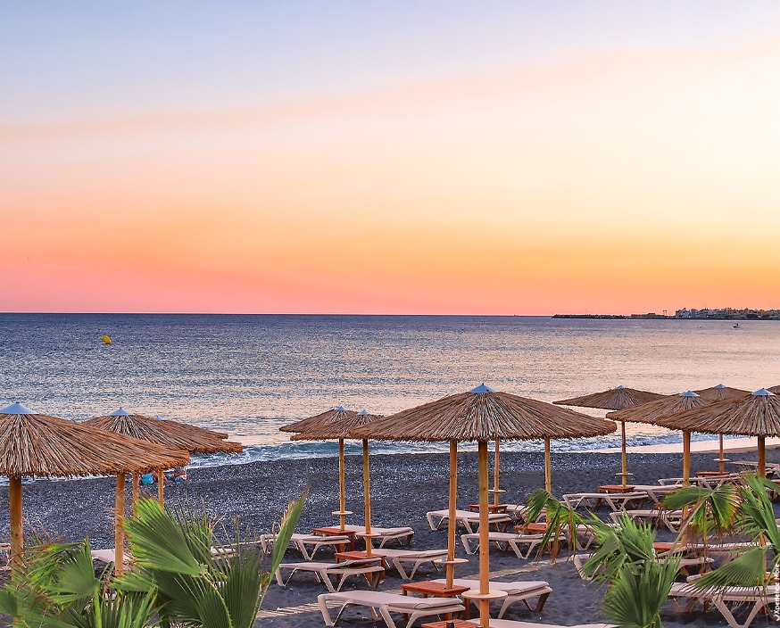 Tylissos Beach Hotel pludmale saulrieta laikā Krētā ar salmu saulessargiem un atpūtas krēsliem pie mierīgas jūras oranžās debesīs