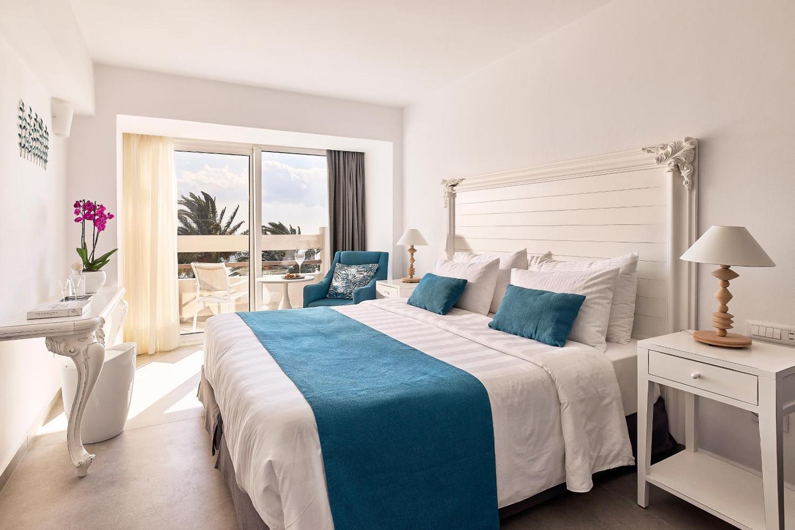 Moderns un gaišs Tylissos Beach Hotel viesnīcas numurs ar baltām mēbelēm, ziliem akcentiem un izeju uz saulainu balkonu.