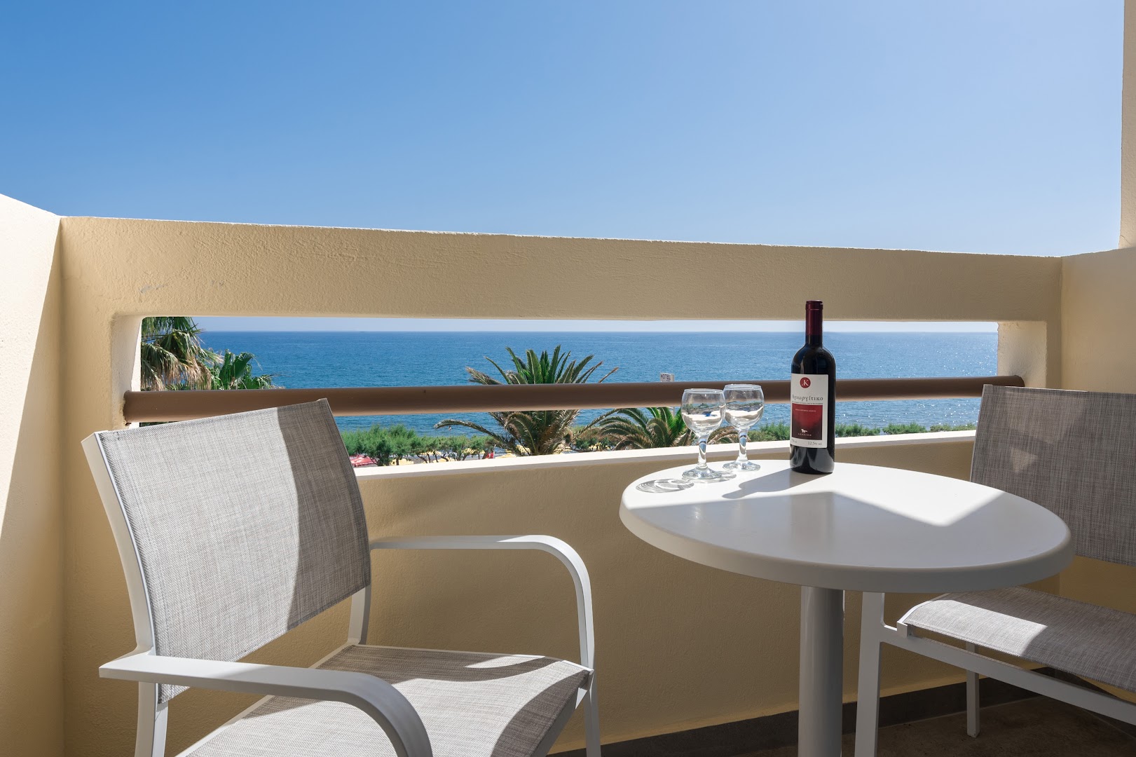 Tylissos Beach Hotel balkons ar galdiņu, krēsliem un vīna pudeli, fonā redzama zilā jūra un piekrastes palmas.