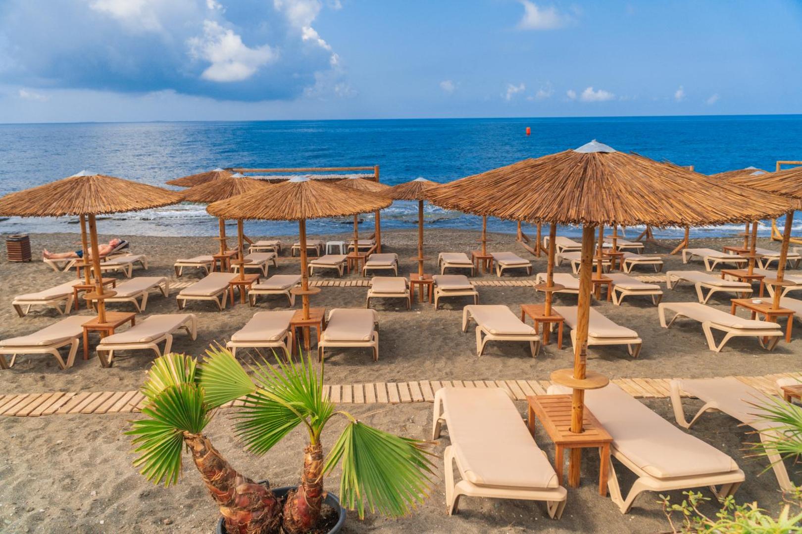 Tylissos Beach Hotel pludmales atpūtas zona ar salmu saulessargiem, ērtiem zviļņiem un koka laipām uz tumšo smilšu fona