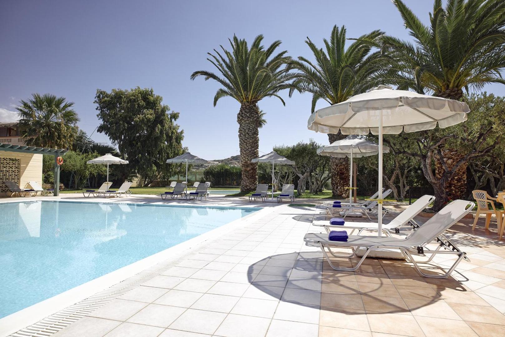 Tylissos Beach Hotel baseina zona dienas vidū — balti saulessargi, ērti zviļņi un augstas palmas zem skaidrām debesīm