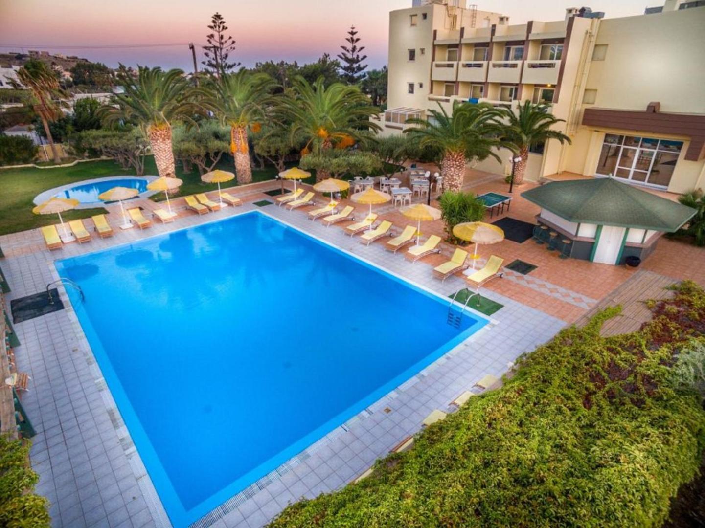 Tylissos Beach Hotel galvenais āra baseins saulrietā — plaša atpūtas zona ar dzelteniem saulessargiem un palmām Krētas piekrastē