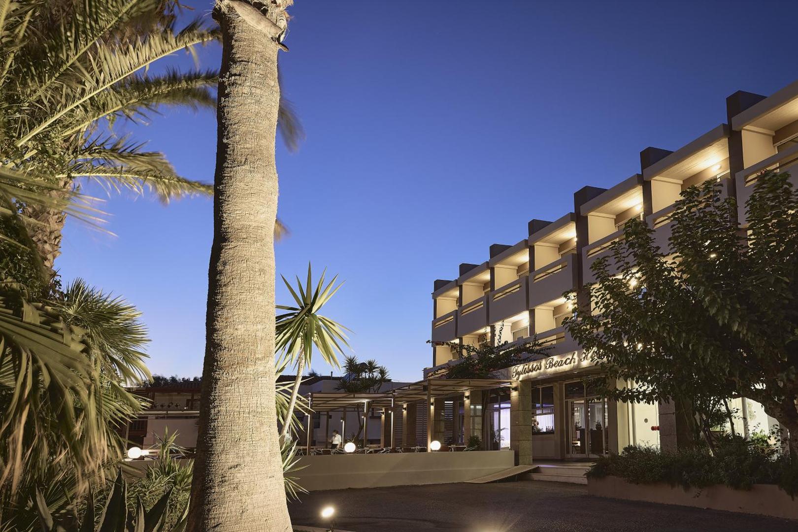 Tylissos Beach Hotel fasāde vakara apgaismojumā ar palmām un āra terasi Krētā — moderna arhitektūra un mājīga atmosfēra