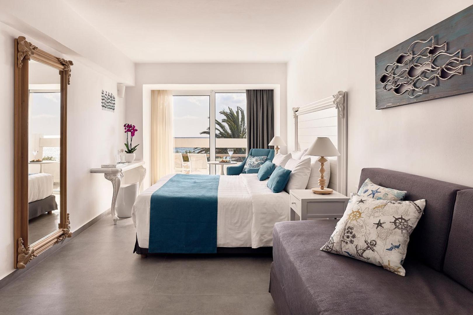 Gaišs Tylissos Beach Hotel numurs ar divguļamo gultu, dīvānu un balkonu, no kura paveras skats uz palmu dārzu.