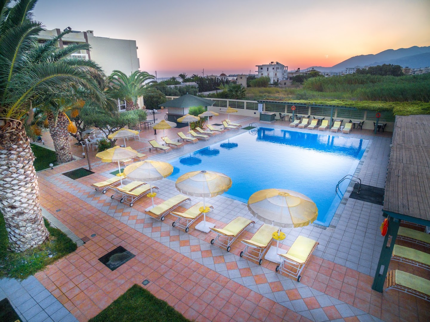 Skats uz Tylissos Beach Hotel baseinu un apkārtnes kalniem saulrieta laikā — atpūtas krēsli un saulessargi sakārtoti pie ūdens