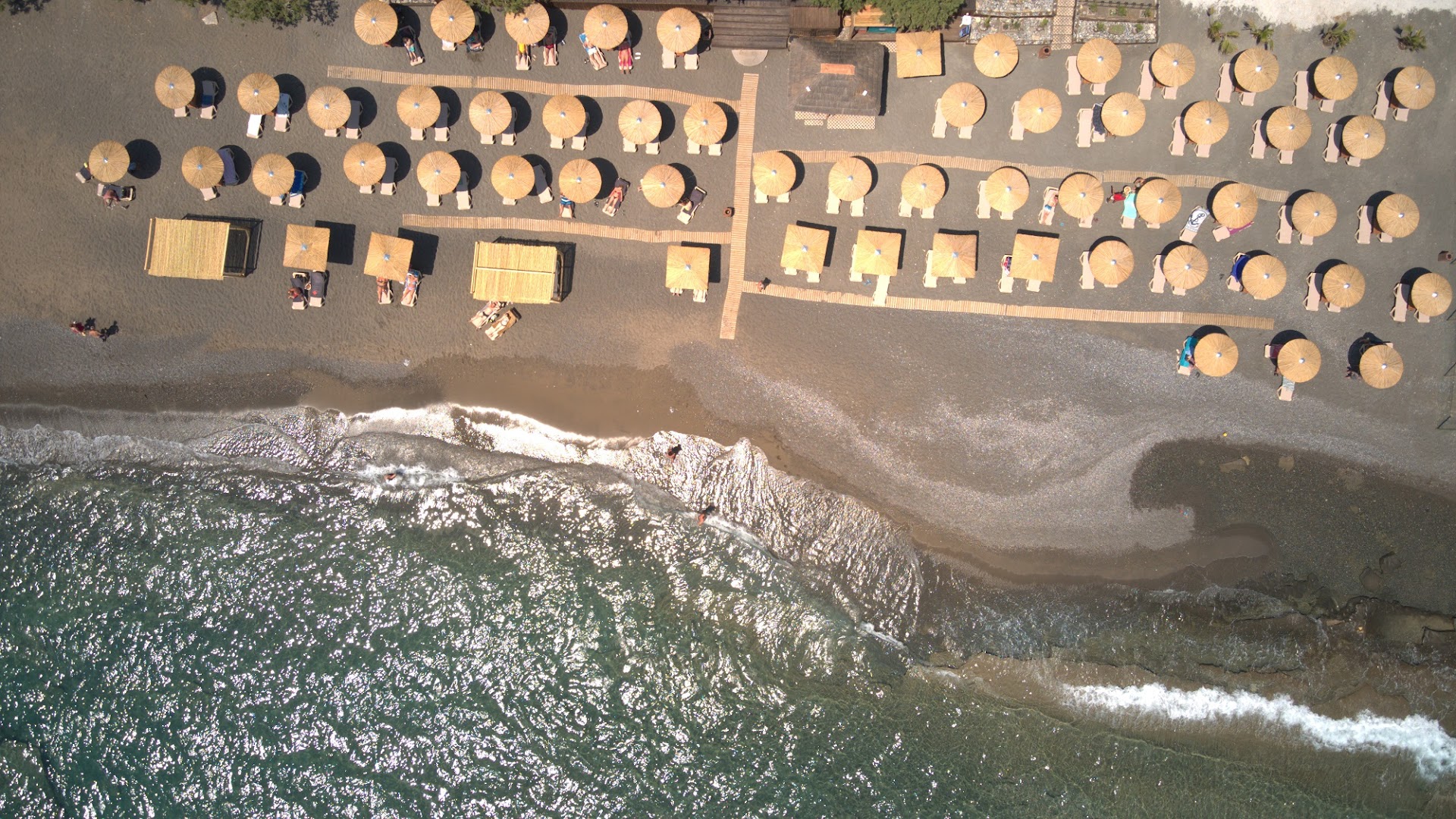 Tylissos Beach Hotel pludmales skats no augšas ar sakārtotām saulessargu rindām, smiltīm un dzidru jūras ūdeni Krētas piekrastē