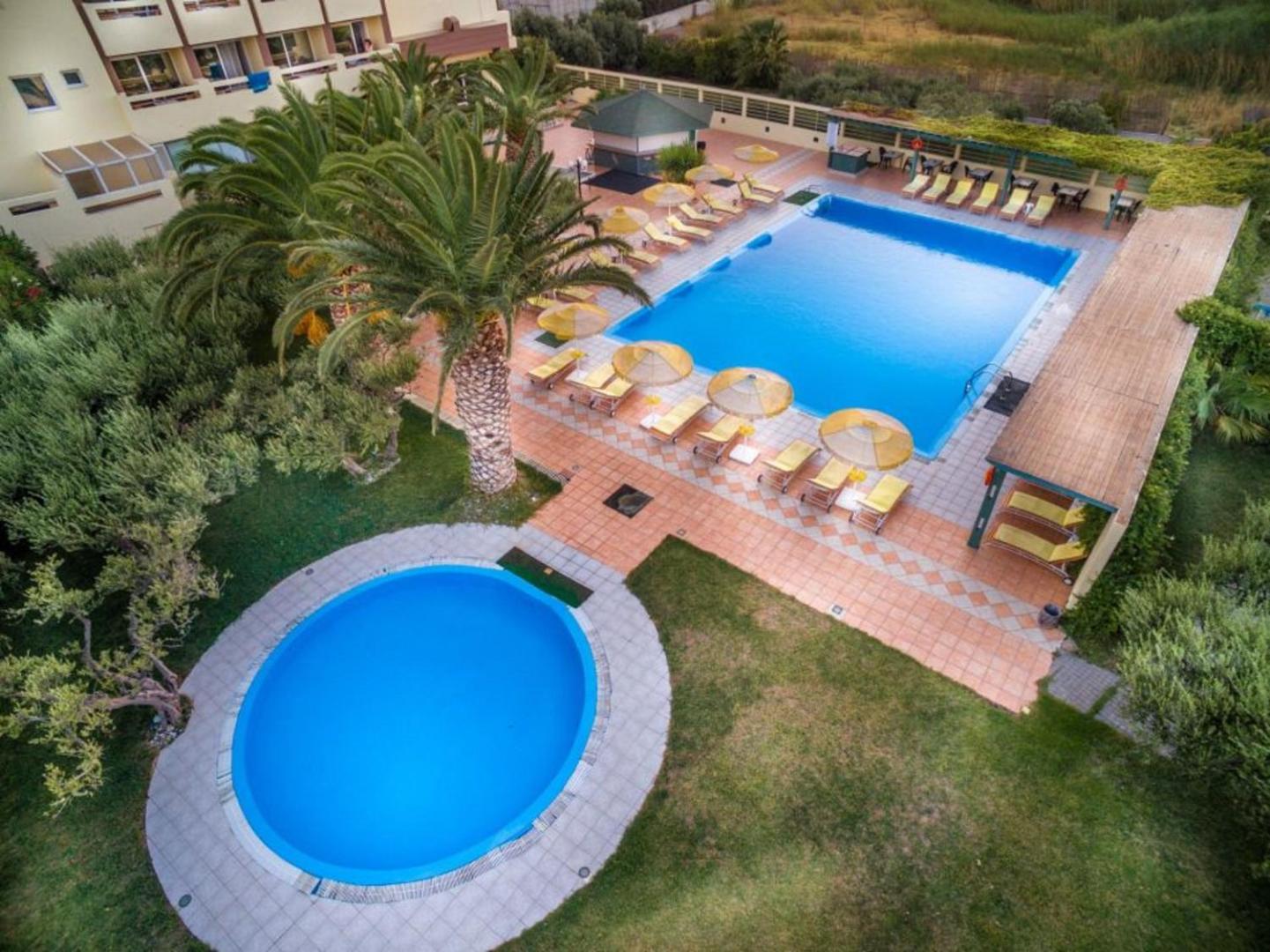 Aerofoto skats uz Tylissos Beach Hotel teritoriju — lielais peldbaseins un apaļais bērnu baseins dārza un palmu ielenkumā
