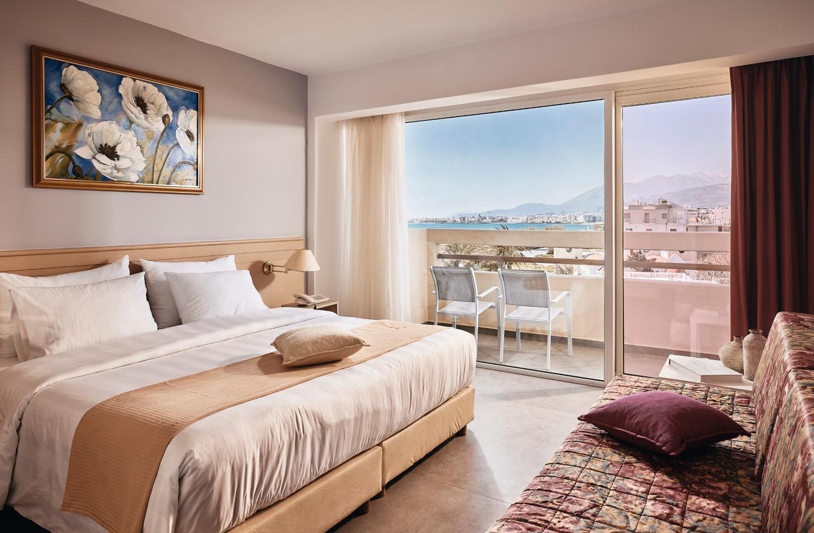 Tylissos Beach Hotel numura interjers ar skatu uz jūru un pilsētu no balkona, aprīkots ar ērtu gultu un dīvānu.