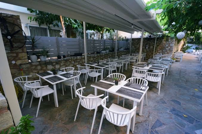 Dimitrios Beach viesnīcas āra terases restorāns ar baltiem krēsliem, koka galdiem un akmens sienu apdari mājīgā atmosfērā